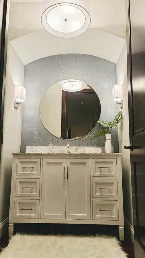 white bathroom vanity with textured wallpaper and large round mirror